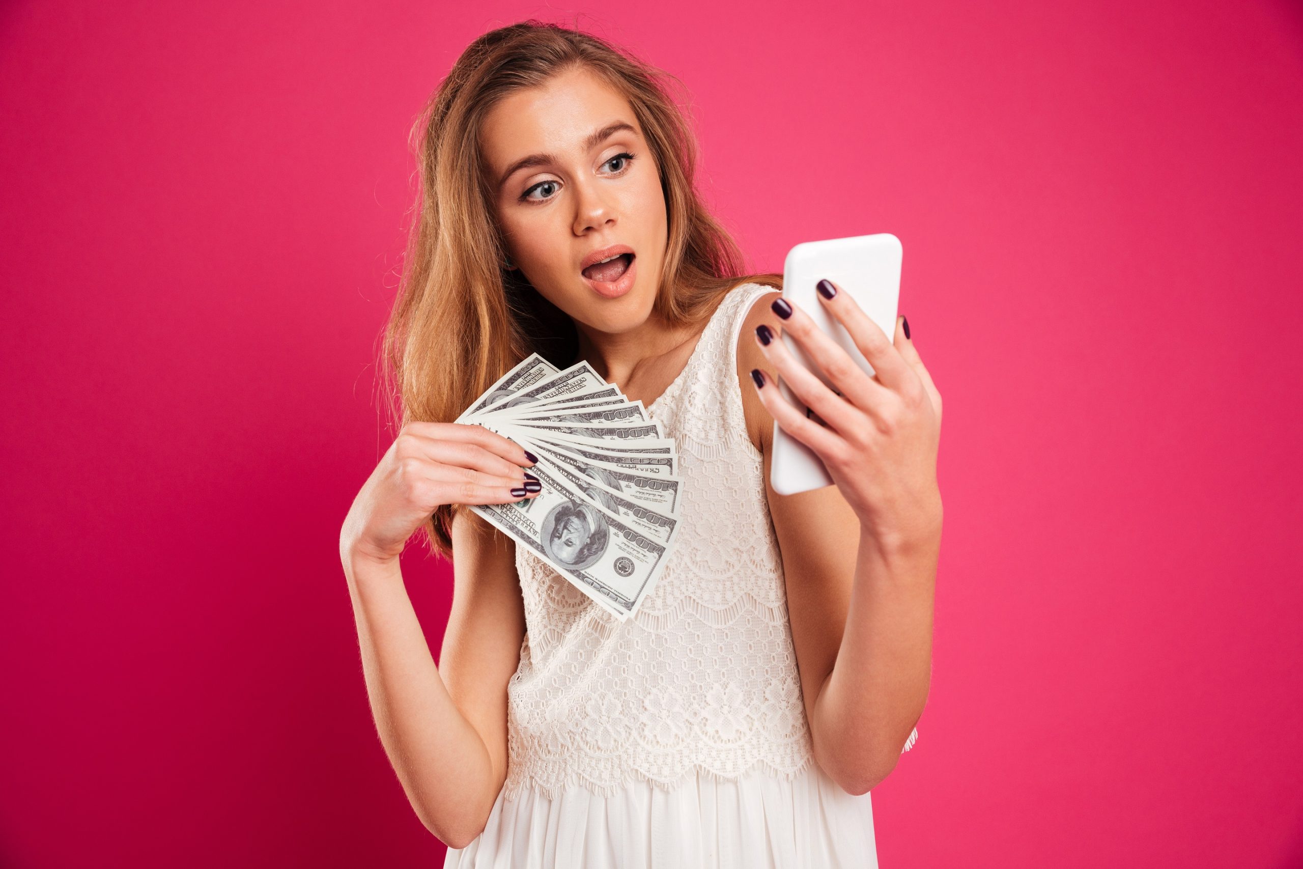 Portrait of a shocked pretty girl holding money banknotes while resding message on mobile phone isolated over pink background